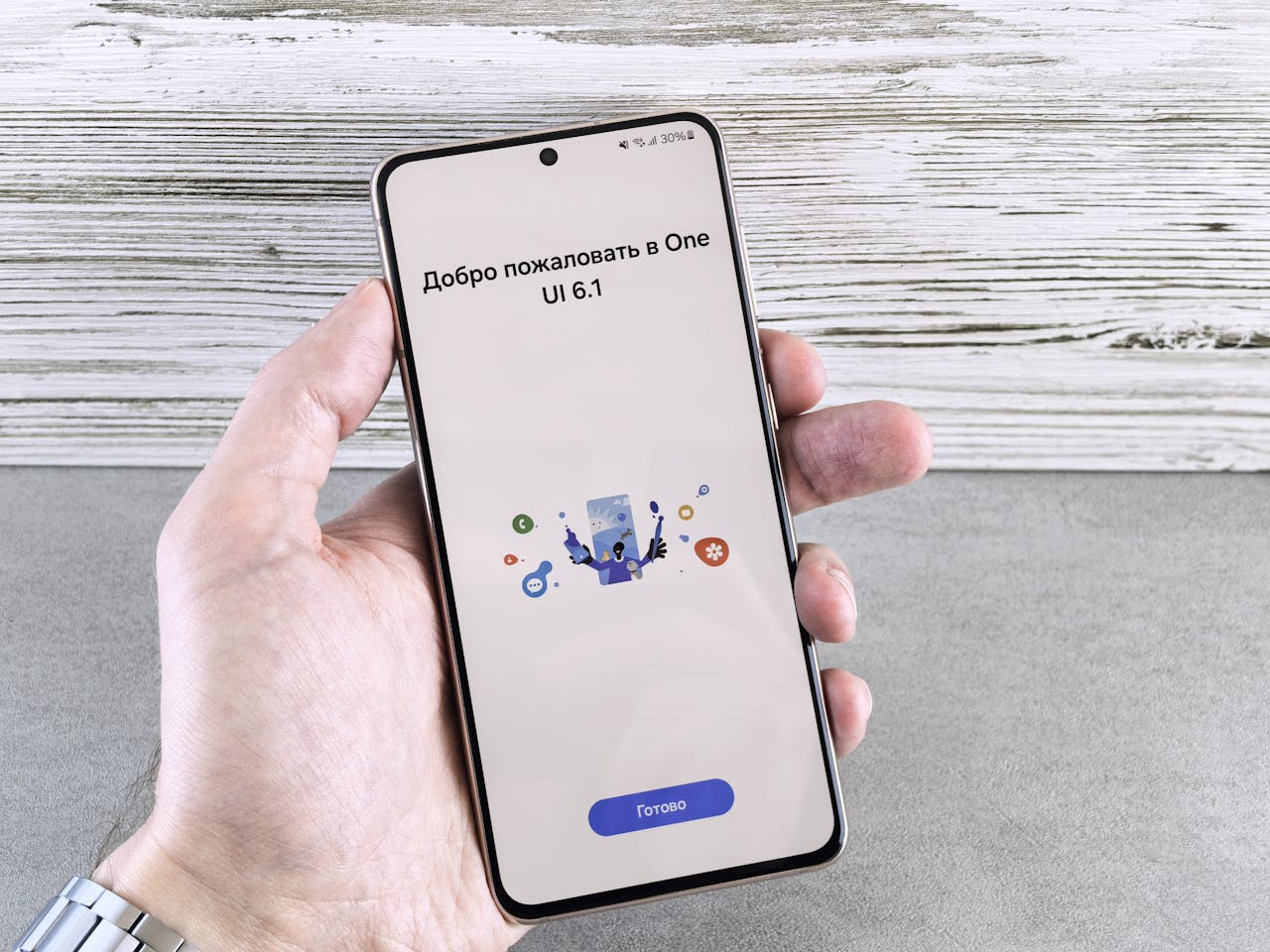 about-03 A close-up of a hand holding a smartphone displaying the One UI 6.1 welcome screen indoors.